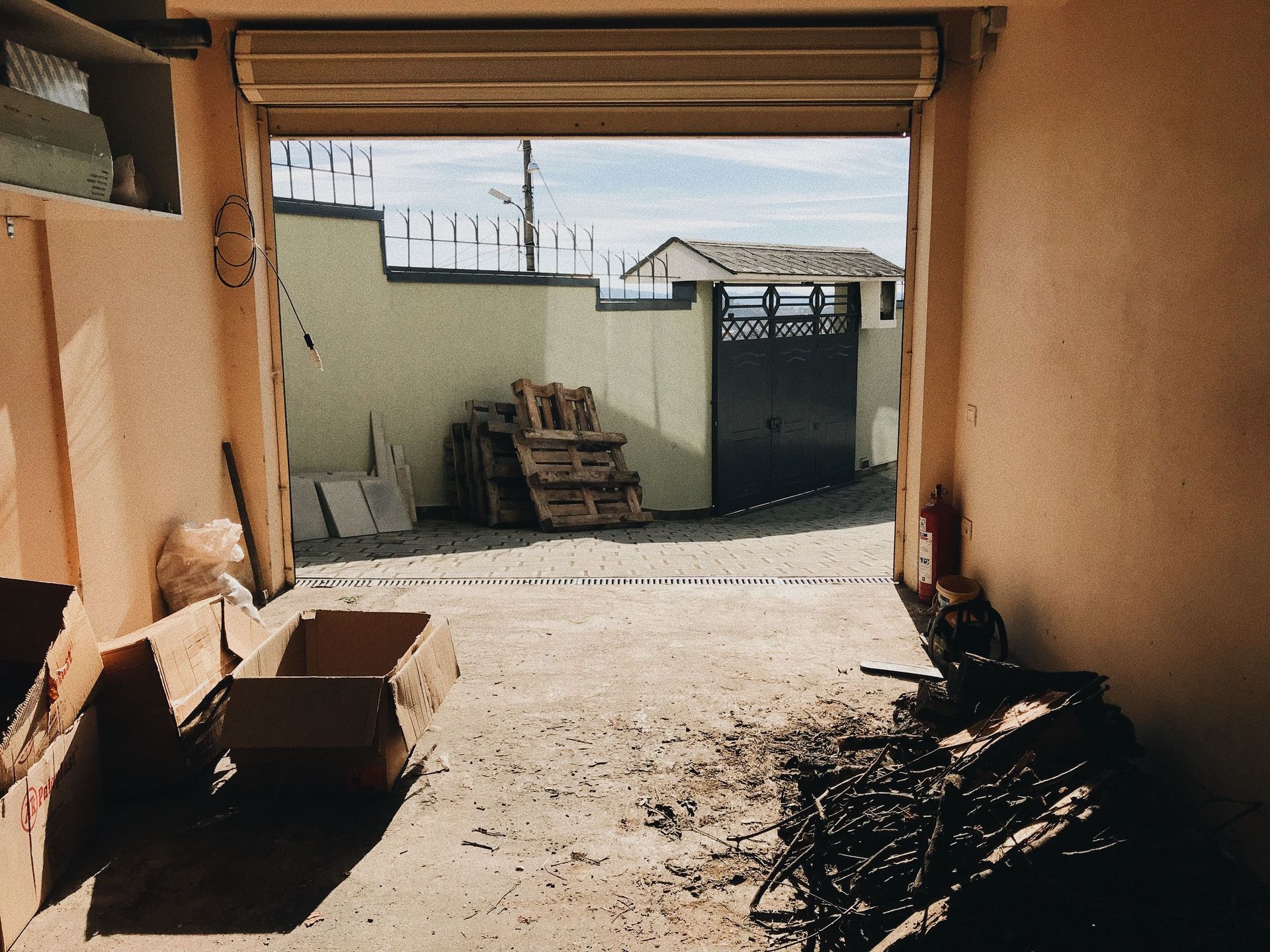 View from inside a garage showing open roller shutter, cardboard boxes, and an outdoor concrete fence.